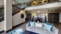 This elegantly designed communal space makes a striking first impression with its dramatic double-height ceilings and open, airy atmosphere. The main lounge area serves as the heart of the space, anchored by a tiled fireplace with a mounted flat-screen television above, flanked by large windows that frame views of the outdoors and flood the room with natural light. The seating area is thoughtfully curated with a plush gray sofa, teal and blue accent pillows, and a geometric-patterned area rug, all arranged around a sleek glass coffee table.

A bold staircase with dark metal railings rises to an upper mezzanine level, which overlooks the lounge below and adds to the sense of vertical grandeur. The space is adorned with distinctive wall art, including a sculptural sunburst installation and abstract canvas paintings, lending it a refined, contemporary character. Pendant and sconce lighting complement the warm, inviting ambiance throughout.

Adjacent to the main lounge, a reception counter with a dark granite surface offers a functional workspace. Nearby, a dedicated fitness center is visible through mirrored glass, equipped with cardio machines. Large-format tile flooring runs consistently throughout, tying the various functional areas together into a cohesive, well-appointed amenity space.