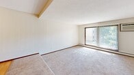 This one-bedroom, one-bathroom unit in Roseville, Minnesota offers a well-organized layout across a single floor. The galley-style kitchen is efficiently designed with rich wood cabinetry on both sides, light countertops, a double sink, and white appliances including a range, refrigerator, and built-in microwave. Light hardwood-style flooring runs through the kitchen, which opens into the combined living area. The living room is bright and carpeted, featuring a ceiling fan with light fixture and a large sliding glass door that leads to a private balcony with outdoor views of mature trees and green surroundings. Vertical blinds frame the sliding door, allowing for natural light control. The spacious bedroom offers plush carpeting, a dedicated window with blinds for natural light, a baseboard heating unit, and a wood-paneled door leading to a closet. The bathroom is clean and functional, featuring a full tiled tub-and-shower surround in crisp white. A hallway connects the main living spaces, providing a logical flow throughout the unit. Textured ceilings and neutral wall tones are consistent throughout, lending a cohesive, understated aesthetic to the overall space.