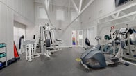 This fitness room is defined by its striking architectural character, most notably the dramatic vaulted ceiling with exposed white-painted timber trusses that lend the space an open, airy quality. White shiplap-style wall paneling and a neutral color palette keep the environment bright and clean, while large windows fitted with shutters allow natural light to filter throughout the room. The durable dark concrete flooring provides a practical, low-maintenance surface suited to active use.

The room is comprehensively outfitted for both cardiovascular and strength training. Cardio equipment includes multiple treadmills, elliptical trainers, and a recumbent stationary bike, offering a range of aerobic workout options. For strength training, a multi-station cable machine and additional resistance equipment are positioned to allow efficient use of the floor space. A rack of kettlebells and free weights provides further versatility for functional fitness routines.

Full-length mirrors line one wall, enhancing the sense of space while allowing users to monitor their form during exercise. A wall-mounted television adds to the amenity, and a ceiling fan aids in air circulation. The room benefits from both a main entry door and an emergency exit, ensuring safe and convenient access. Overall, this fitness room combines practical functionality with a bright, well-designed environment suitable for a complete workout experience.