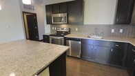 This single-floor kitchen is thoughtfully designed with a cohesive blend of dark espresso cabinetry and light granite countertops, creating a striking contrast that anchors the space. The full suite of stainless steel appliances—including an electric range, over-the-range microwave, and dishwasher—provides both functionality and a polished, contemporary aesthetic. A neutral gray rectangular tile backsplash runs the length of the cooking and prep area, adding texture while complementing the overall color palette. The kitchen features a large island with an expansive granite surface, offering generous additional workspace and the potential for casual seating or entertaining. Dark hardwood flooring runs throughout, reinforcing the modern tone of the space. The under-cabinet layout maximizes storage with upper and lower cabinets flanking the appliances, while the sink area is positioned along the far counter for practical workflow. An entryway is visible adjacent to the kitchen, hinting at an open, connected floor plan. The space balances style and utility effectively.