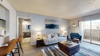 This thoughtfully designed single-story residence offers 1 bedroom and 2 bathrooms across a compact yet efficient floor plan. The living room serves as the heart of the home, featuring wood-style flooring, a textured ceiling, and a bold blue accent wall adorned with round decorative mirrors. Sliding glass doors open directly to a private balcony, filling the space with natural light. The dining area continues the blue accent wall theme, creating a cohesive and stylish aesthetic throughout the common areas. The kitchen is efficiently laid out with white cabinetry, light countertops, a black electric range, and a dishwasher, maximizing functionality within the available space. The spacious primary bedroom features plush carpet flooring, a large window for natural light, and generous wall space. A dedicated walk-in closet with multiple hanging rods and built-in shelving provides ample storage. The two bathrooms include a tub-shower combination with clean white tile surround. Wood-style flooring runs through the main living areas, complementing the neutral gray walls and modern finishes seen throughout. The overall ambiance is contemporary and inviting, with a cohesive blue and neutral color palette tying each room together.