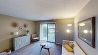 This single-floor residence offers a thoughtfully arranged layout connecting living, dining, kitchen, and sleeping spaces through wood-look flooring and a central hallway. The living room is carpeted and centers around a large sliding glass door fitted with vertical blinds, allowing natural light to filter in while providing outdoor access and views of greenery. Decorative wall accents and a console cabinet add character to the space. Adjacent to the living room, the dining area accommodates a glass-top table with patterned upholstered chairs beneath a ceiling fan with integrated lighting. A doorway connects directly to other areas of the residence. The galley-style kitchen features dark espresso cabinetry, granite-look countertops, a double-basin sink, and black appliances including a refrigerator and electric range, all set over wood-look flooring. The bedroom is carpeted, with natural light provided by a window fitted with horizontal blinds and space for bedside furnishings. A hallway with wood-look flooring and a thermostat connects the rooms to the bathroom, which includes a dark vanity with granite countertop, a large mirror, and a lighted vanity fixture above.