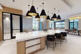 Located at 2120 Dunavant St in Charlotte's vibrant South End neighborhood, this elegantly designed single-floor amenity space offers a seamless blend of modern aesthetics and functional living. The open-concept layout is unified by large-format porcelain tile flooring that flows throughout, creating a bright and cohesive environment.

The centerpiece of the space is a striking kitchen area anchored by an expansive white quartz island with bar seating, topped by a dramatic cluster of black-and-gold conical pendant lights. Warm wood cabinetry, integrated appliances, and a decorative patterned accent wall lend the kitchen area a refined yet inviting character.

The living lounge area is equally impressive, featuring a tray ceiling with integrated cove lighting and a sculptural gold ring chandelier that adds a bold artistic focal point. A generous sectional sofa and curated seating arrangements create comfortable conversation areas, while floor-to-ceiling windows and glass doors flood the space with natural light and provide views of the surrounding streetscape and greenery.

Structural columns clad in natural stone add an architectural dimension throughout, and interior glass partitions define distinct zones while maintaining visual openness. The overall ambiance is contemporary and upscale, with a warm neutral palette, carefully chosen furnishings, and thoughtful lighting that together create an inviting and polished environment.