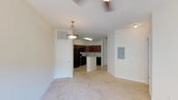 This single-floor, one-bedroom, one-bathroom unit features an open-concept layout that seamlessly connects the living, dining, and kitchen areas. The living area is finished with light-toned carpeting, creating a warm and neutral foundation throughout the space. A ceiling fan overhead provides both comfort and functionality.

The kitchen is positioned toward the rear of the open floor plan and is equipped with dark cabinetry, black appliances including a refrigerator and range, and a peninsula that provides additional counter space and serves as a natural divider between the kitchen and the living and dining areas. A pendant light hangs above the peninsula, adding a focused decorative touch. The kitchen also features tile or hard flooring, contrasting with the carpeted living area.

A white paneled door near the kitchen provides access to additional storage or a closet. An electrical panel is mounted on one wall, and HVAC vents are positioned in the ceiling. The overall palette is light and neutral, with white walls and trim contributing to a clean, open ambiance throughout the unit.