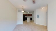 This single-floor, one-bedroom, one-bathroom unit features an open-concept layout that seamlessly connects the living, dining, and kitchen areas. The living area is finished with light-toned carpeting, creating a warm and neutral foundation throughout the space. A ceiling fan overhead provides both comfort and functionality.

The kitchen is positioned toward the rear of the open floor plan and is equipped with dark cabinetry, black appliances including a refrigerator and range, and a peninsula that provides additional counter space and serves as a natural divider between the kitchen and the living and dining areas. A pendant light hangs above the peninsula, adding a focused decorative touch. The kitchen also features tile or hard flooring, contrasting with the carpeted living area.

A white paneled door near the kitchen provides access to additional storage or a closet. An electrical panel is mounted on one wall, and HVAC vents are positioned in the ceiling. The overall palette is light and neutral, with white walls and trim contributing to a clean, open ambiance throughout the unit.