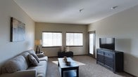 This single-floor space offers a comfortable and well-appointed living area designed with a clean, contemporary aesthetic. The living room serves as the central gathering space, featuring plush carpeted flooring in a warm neutral tone that complements the soft beige walls. A large window arrangement along one wall allows generous amounts of natural light to fill the room, creating an airy and welcoming atmosphere throughout the day. An additional door with a glass panel provides further access to natural light and views of the outdoors. The living area is thoughtfully laid out to accommodate both relaxed seating and entertainment, with ample space for a sofa arrangement and a media console. Dark-accented furniture pieces, including a media cabinet and coffee table, contrast tastefully against the lighter tones of the walls and flooring. A ceiling-mounted fixture provides overhead lighting, while a floor lamp adds a warm, ambient glow to the corner of the room. The overall design is cohesive and understated, with dark baseboards providing a subtle architectural detail that frames the space. The single-floor layout ensures easy accessibility and a straightforward flow, making this a practical and inviting living environment suited for everyday comfort.