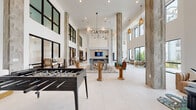 This contemporary clubhouse in Pooler, Georgia, is designed as a dynamic social hub, characterized by its dramatic double-height ceilings, crisp white walls, and large black-framed windows that flood the interior with natural light and offer views of the surrounding community. Weathered wood-clad structural columns rise throughout the expansive main hall, adding warmth and texture against the otherwise modern, light-toned tile flooring.

The central gathering area serves as the heart of the space, anchored by a full-size pool table atop a geometric patterned area rug, alongside a foosball table and a shuffleboard table — catering to a range of recreational activities. Sculptural, eclectic furniture pieces including uniquely crafted wooden chairs are distributed throughout, lending an artistic character to the environment.

A built-in bar area features a bold blue island with woven barstools, white countertops, and a textured tile backsplash beneath a wall-mounted television — ideal for casual gatherings. A separate counter-height media nook with green upholstered stools provides additional seating. Intimate lounge corners with comfortable chairs and floor-to-ceiling windows invite quiet relaxation, while dining-style tables with natural wood chairs offer flexible seating throughout. Pendant lighting and ring chandeliers add refined finishing touches to this well-appointed communal space.