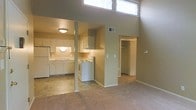This single-floor space is defined by its impressively tall ceilings, which lend a sense of openness and volume well beyond its footprint. High clerestory windows positioned near the ceiling's peak allow natural light to filter into the main living area, brightening the neutral, warm-toned walls throughout.

The open-concept layout connects the living area directly to the kitchen, which is equipped with white cabinetry, a double-basin sink, a white electric range, and a full-size refrigerator. The kitchen features tile flooring, under-cabinet lighting, and a ventilation hood above the stove, providing a practical and well-lit cooking environment. A structural column marks the boundary between the kitchen and living spaces without fully closing off either area.

The carpeted living area offers a comfortable, cohesive feel and flows toward a hallway leading to additional rooms. A doorway visible from the main space opens to a walk-in closet, suggesting a bedroom nearby. A wall-mounted thermostat and a visible HVAC vent indicate climate control throughout. The overall ambiance is clean, neutral, and functional, with the dramatic ceiling height serving as the space's most notable architectural feature.