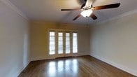 This single-floor space showcases a well-appointed interior that balances classic architectural details with functional design. The main living area is anchored by rich hardwood floors that reflect natural light, creating a warm and inviting atmosphere throughout. Crown molding runs along the perimeter of the ceiling, adding a refined, traditional character to the space.

A prominent set of French doors serves as a central architectural feature, framed by white trim and fitted with slatted blinds for privacy control. These doors allow abundant natural light to filter into the room while providing access to the exterior. The surrounding windows further enhance the brightness of the interior, offering views of the outdoors.

The ceiling features a classic five-blade ceiling fan with an integrated light fixture, providing both ambient lighting and air circulation. The walls are finished in a neutral tone that complements the warm hues of the hardwood flooring, contributing to an overall cohesive and understated aesthetic. The combination of traditional molding, quality flooring, and well-placed openings gives this space a timeless, character-rich quality suited to a variety of uses.