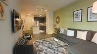 This single-floor space showcases an open-concept layout that seamlessly connects the living area and kitchen, creating a cohesive and functional environment. The living room is anchored by a plush gray sofa, a geometric-patterned area rug, and a glass-top coffee table, all complementing the space's modern character. A sage green accent wall adds a pop of color, enhancing the warm ambiance alongside framed wall art and a decorative sunburst mirror. Track lighting runs along the ceiling, providing adjustable illumination throughout the space, while recessed lights and a pendant light over the kitchen add layered lighting options. The kitchen features dark cabinetry, stainless steel appliances including a refrigerator and microwave, and a peninsula with bar-height green stools that serve as a casual dining and entertaining area. The dark hardwood-style flooring flows continuously from the living area into the kitchen, unifying the space visually. The entry door is visible near the kitchen, and the overall layout is efficient and well-organized, making thoughtful use of the available footprint.