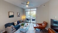 This single-floor residence at 6806 Morrison Blvd in Charlotte, NC offers a thoughtfully designed open-concept layout with contemporary finishes throughout. The kitchen is a standout feature, showcasing sleek white cabinetry with a high-gloss finish, a green glass tile backsplash, stainless steel appliances including a French door refrigerator and range, and a generous quartz peninsula with seating for multiple guests. Pendant lighting above the peninsula and recessed ceiling lights provide layered illumination, while hardwood-style flooring flows throughout the main living areas.

The living room is bright and inviting, benefiting from a ceiling fan and large floor-to-ceiling sliding glass doors that open onto a private balcony, offering outdoor seating and views of surrounding greenery. The bedroom features plush carpeting, a ceiling fan, angled architectural ceiling details, and a window allowing natural light to filter in. Neutral wall tones throughout create a calm, cohesive atmosphere. The bathroom includes tile flooring, a tub-shower combination with white subway tile surround, a vanity with quartz countertop, and a lighted mirror. Overall, the space presents a clean, modern aesthetic well-suited to comfortable everyday living.