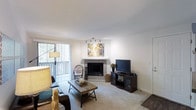 This single-floor residence offers a welcoming and well-appointed layout designed for comfortable everyday living. The living room serves as the heart of the space, centered around a tiled corner fireplace that adds warmth and a focal point to the room. Soft, neutral-toned carpet flooring extends throughout, complementing the crisp white walls and creating a clean, inviting atmosphere. A sliding glass door floods the room with natural light while offering views of the outdoor greenery beyond, enhanced by vertical blinds for adjustable privacy. Track lighting on the ceiling provides additional illumination, ensuring the space is well-lit at all times. The living area is thoughtfully arranged with a mix of seating, including a wicker accent chair and cushioned sofa, gathered around a rustic wooden coffee table. A media console with built-in storage sits adjacent to the fireplace, offering a practical entertainment setup. Colorful wall art above the mantle adds a vibrant artistic touch to the otherwise neutral palette. The overall ambiance is warm, relaxed, and tastefully styled, making it a comfortable and functional single-floor living space.