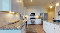 This single-floor residence showcases a contemporary design with a thoughtful, open layout connecting the primary living spaces. The kitchen serves as a central highlight, outfitted with crisp white shaker cabinetry, granite countertops, a classic white subway tile backsplash, and a full suite of stainless steel appliances including a gas range, over-the-range microwave, and refrigerator. A central island with seating for two is illuminated by pendant lighting with brass-toned fixtures, making it ideal for casual dining or entertaining. Light wood-tone flooring runs throughout the kitchen and living areas, enhancing the bright, airy atmosphere.

The living room benefits from generous natural light through large windows dressed with bold patterned curtains, creating a warm and inviting gathering space. Two well-sized bedrooms offer plush carpeting, oversized windows, and ample room for furnishings. Both bathrooms are finished with white cabinetry, granite countertops, large vanity mirrors, and white tile flooring, with one featuring a walk-in glass shower and the other a shower-tub combination.