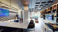 The club room at 55 N Water St in Norwalk, Connecticut, is a thoughtfully designed communal space that blends sophisticated aesthetics with practical amenities. The room is defined by its striking coffered ceiling finished in deep charcoal, paired with warm herringbone-patterned wood-look flooring that creates a refined contrast throughout the space.

The lounge area anchors the room with classic leather Eames-style lounge chairs, marble-topped accent tables, and a working fireplace framed by built-in shelving with gold-accented cabinetry. The shelving wall also houses a large flat-screen television, making it an ideal space for both relaxed viewing and socializing. A dedicated game table adds an interactive element to this area.

A long white quartz countertop peninsula serves as a bar-style gathering space, lined with sleek black and gold barstools beneath a cluster of brass pendant lights. Adjacent to this is a fully equipped kitchenette with navy cabinetry, white countertops, a coffee station, microwave, and refrigerator.

Floor-to-ceiling windows at one end of the room draw in natural light and offer outdoor views, while recessed and wall-mounted lighting adds warmth throughout. The overall ambiance is modern, upscale, and inviting.