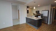 This contemporary single-floor space showcases a thoughtfully designed open-concept layout that seamlessly blends style and functionality. The kitchen serves as the centerpiece of the home, featuring a large island topped with granite countertops and an integrated sink, flanked by a built-in wine rack for added character. Dark cabinetry on the island contrasts elegantly with crisp white upper cabinets, creating a bold two-tone aesthetic. Stainless steel appliances, including a French door refrigerator, electric range, and microwave, complete the modern kitchen ensemble. Pendant lighting in mixed finishes hangs above the island, providing both task lighting and visual interest. Warm hardwood-style flooring flows throughout, adding a welcoming and cohesive feel to the space. Recessed lighting and a clean, neutral color palette on the walls contribute to a bright, airy ambiance. A sliding barn-style door provides a stylish transition to an adjacent room, visible in the background, which features carpeted flooring. The overall design strikes a balance between contemporary urban aesthetics and comfortable everyday living.
