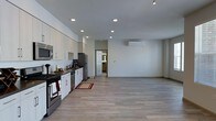 This single-floor space presents a clean, modern design centered around an open-concept floor plan that seamlessly connects the kitchen and living areas. Light-toned hardwood-style flooring runs throughout, contributing to a bright and cohesive aesthetic.

The kitchen is well-appointed with white cabinetry, dark stone countertops, and a full suite of stainless steel appliances, including a gas range, refrigerator, and built-in microwave. Recessed ceiling lights and a dedicated track lighting fixture provide ample, adjustable illumination across the space.

The expansive living area offers generous room for a variety of furniture configurations. Large windows fitted with horizontal blinds allow natural light to fill the room while providing privacy control. A wall-mounted mini-split HVAC unit ensures year-round climate comfort without the need for traditional ductwork. The neutral wall tones and smooth ceilings create a minimalist backdrop that is both versatile and inviting.

A doorway visible from the main living area leads to an additional room, suggesting further private or functional space beyond the open layout. Overall, the space combines contemporary finishes with practical design, making it a functional and visually appealing environment.