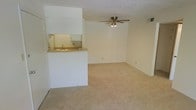 This single-floor space features an open-concept design that connects the kitchen and living area in a functional, straightforward layout. The living area is finished with wall-to-wall carpeting in a neutral beige tone, contributing to a clean and understated aesthetic. A ceiling fan with a built-in light fixture is centrally positioned overhead, providing both air circulation and ambient lighting. The textured ceiling adds a subtle dimensional quality to the space.

The kitchen is partially separated from the living area by a white half-wall partition, which incorporates a pass-through counter with a sink. This design element maintains an open sightline between the two areas while defining each zone. Upper cabinetry is visible above the pass-through, offering storage space, and the cabinetry continues within the kitchen, finished in white to match the surrounding walls. A mirrored backsplash reflects light within the kitchen area.

The walls throughout are painted in a light, neutral tone, lending the space a bright and airy feel. A wall vent is visible near the ceiling, indicating a built-in HVAC system. Overall, the space presents a simple, functional interior with consistent finishes.