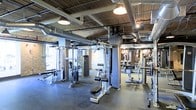 This fitness facility offers a comprehensive workout environment designed to accommodate a wide range of training needs. The main floor is generously sized, providing ample space for members to move freely between stations without congestion. The layout is thoughtfully organized, with a robust selection of strength training cable machines and weight equipment arranged throughout the floor, allowing for efficient circuit-style training or focused muscle group workouts.

The interior architecture lends the space a distinctly industrial character. Exposed brick columns punctuate the floor, while overhead, bare ductwork and structural beams run across the ceiling alongside warm pendant lighting and track-style fixtures. This raw aesthetic is complemented by rubber flooring underfoot, which provides both durability and safety during workouts.

Large windows along one wall admit natural light, brightening the space and creating a more open, energizing atmosphere. Mirrored walls on the opposite end visually expand the room while allowing members to monitor their form. Motivational imagery adorns the brick walls, contributing to the facility's focused and driven ambiance. Overall, this gym combines functional equipment density with an industrial aesthetic to create a practical and motivating training environment.
