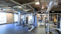 This fitness facility offers a comprehensive workout environment designed to accommodate a wide range of training needs. The main floor is generously sized, providing ample space for members to move freely between stations without congestion. The layout is thoughtfully organized, with a robust selection of strength training cable machines and weight equipment arranged throughout the floor, allowing for efficient circuit-style training or focused muscle group workouts.

The interior architecture lends the space a distinctly industrial character. Exposed brick columns punctuate the floor, while overhead, bare ductwork and structural beams run across the ceiling alongside warm pendant lighting and track-style fixtures. This raw aesthetic is complemented by rubber flooring underfoot, which provides both durability and safety during workouts.

Large windows along one wall admit natural light, brightening the space and creating a more open, energizing atmosphere. Mirrored walls on the opposite end visually expand the room while allowing members to monitor their form. Motivational imagery adorns the brick walls, contributing to the facility's focused and driven ambiance. Overall, this gym combines functional equipment density with an industrial aesthetic to create a practical and motivating training environment.
