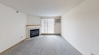 This single-floor residential unit offers a practical and functional layout designed for comfortable everyday living. The open-concept living area is a central highlight, featuring plush carpeting, a tile-surround fireplace, and a sliding glass door that opens to a balcony, allowing natural light to fill the space. A ceiling fan with an integrated light fixture adds both comfort and style to the main living area. The kitchen is efficiently designed with white cabinetry, a stainless steel refrigerator, black appliances including a built-in oven and microwave, a tile floor, and a convenient pass-through counter that connects to the living space. The bedrooms are carpeted and well-lit, each equipped with baseboard heating and wall-unit air conditioning, with select rooms offering sliding glass doors or windows with outdoor views. Two bathrooms serve the unit — one featuring a full tub-shower combination with clean white tile surround, and a second with a standalone glass-enclosed shower stall and tile flooring. Additional storage is accessible via closets with wire shelving. Textured ceilings are consistent throughout the unit, complementing the neutral white walls and warm wood-toned trim that unify the overall aesthetic.