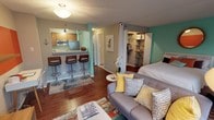 This thoughtfully designed studio offers a smart, open-concept floor plan that maximizes every inch of its single-level layout. Rich hardwood flooring flows throughout, tying together the living, sleeping, and dining areas into one cohesive space. A bold teal accent wall anchors the sleeping area, which is defined by floor-to-ceiling curtain panels that offer a sense of separation without sacrificing openness. A well-organized walk-in closet with built-in shelving sits just beside the sleeping zone, providing ample storage.

The kitchen is positioned along one side, featuring warm wood cabinetry, granite-style countertops, a double-basin sink, and a black electric range. A raised breakfast bar with bar stool seating serves as both a dining area and a natural divider between the kitchen and the main living space. An accent wall painted in bold red-orange adds a dynamic visual element to the living area, complemented by wall-mounted shelving.

Large windows frame striking high-rise urban views, filling the space with natural light. The bathroom includes a wood-framed vanity mirror, granite-topped cabinetry, and a full tub-shower combination. The overall ambiance is colorful, modern, and highly functional.