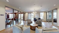 This single-floor community space is thoughtfully designed to offer a range of comfortable, functional areas suited for both relaxation and social gatherings. The main lounge area greets visitors with elegant chandelier lighting, recessed ceiling details with exposed wooden beams, and a sleek linear fireplace flanked by wall-mounted media. Plush seating arrangements, neutral-toned textured wallcovering, and warm tile flooring create an inviting and polished ambiance throughout. Framed artwork and carefully selected accent lighting add character to the space.

Adjacent to the lounge, a bright reception and seating area features large windows that flood the room with natural light and offer views of the surrounding landscape. Glass partition walls with sliding doors connect the various zones, promoting an open, flowing layout while maintaining distinct areas for different activities.

The expansive dining and event room accommodates multiple seating configurations around solid wood tables and modern swivel chairs, complemented by a coffered ceiling with wood beam accents and a wall-mounted television. A kitchen area with stainless steel appliances supports catering and event needs.

Sliding glass doors open to a covered outdoor terrace furnished with patio dining sets, offering scenic open-area views — an ideal extension of the interior gathering space.
