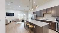 This single-floor open-concept space seamlessly combines a fully equipped kitchen with a comfortable living area, creating a bright and inviting communal environment. The kitchen is a standout feature, anchored by a large granite-topped island with bar seating for multiple guests, making it ideal for casual dining and social gatherings. Dark espresso cabinetry extends to the ceiling, offering generous storage, while a textured geometric tile backsplash adds visual interest. Stainless steel appliances — including a French door refrigerator with ice maker, microwave, and a built-in beverage cooler — complete the well-appointed cooking space. Three industrial-style pendant lights hang above the island, providing focused task lighting with a contemporary flair. The living area transitions naturally from the kitchen, featuring high ceilings that lend a sense of openness. Large windows and glass French doors flood the space with natural light and offer direct views of an outdoor pool and landscaped grounds. Light hardwood-style flooring flows consistently throughout, unifying the space. The lounge area is furnished with upholstered armchairs and a wall-mounted flat-screen TV, while a narrow console table and bar stools along one wall provide additional flexible seating. Abstract artwork adds a finishing touch of personality to the clean, neutral palette.