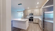 This single-floor, 3-bedroom, 2-bathroom residence offers a thoughtfully designed layout with modern finishes throughout. The open-concept kitchen and living area serves as the heart of the home, anchored by a large central island with a granite countertop and an integrated sink, complemented by light wood cabinetry extending to the ceiling for ample storage. Stainless steel appliances, including a range, microwave, and refrigerator, complete the well-equipped kitchen. Light wood-look flooring flows through the main living areas, adding warmth and continuity to the space. The living room benefits from natural light streaming through large windows and a sliding glass door that opens to a private outdoor balcony, with a ceiling fan adding comfort overhead. The three bedrooms feature plush wall-to-wall carpeting and neutral tones, with the secondary bedrooms offering bright windows with outdoor views. Both bathrooms are finished with light wood vanities, granite countertops, and large-format tile flooring. One bathroom features a soaking tub with tiled surround, while the other includes an elongated vanity with generous storage. Clean lines, recessed lighting, and a neutral color palette throughout give this residence a fresh, contemporary feel.