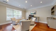 Located at 1200 Somerby Dr in Mobile, Alabama, this single-room living space offers a thoughtfully designed and elegantly finished interior. Rich, warm-toned hardwood floors run throughout, providing a striking foundation that complements the room's neutral, soft beige walls. Crown molding lines the ceiling perimeter, adding a refined architectural detail that enhances the overall sense of polish and quality.

Recessed lighting is distributed evenly across the ceiling, ensuring consistent illumination throughout the space. Multiple windows fitted with white horizontal blinds allow abundant natural light to filter in while offering privacy control, and views of lush greenery outdoors contribute a calm, serene atmosphere.

The living area is arranged with comfortable upholstered armchairs surrounding a light blue tufted ottoman, centered on a neutral area rug. A decorative media console with quatrefoil-patterned cabinet doors houses a flat-screen television and is flanked by framed artwork, adding visual character to the space.

A built-in counter workspace with a granite-style surface and cabinetry below provides a functional desk area, complete with bar-height seating — ideal for productivity within the living environment. A small wet bar with a sink and open shelving is also present, enhancing the room's utility. A glass-paneled entry door connects the space to the exterior, further brightening the interior.