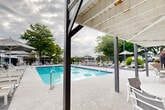 Located at 1011 Gadd Rd in Chattanooga, Tennessee, this outdoor pool area is a well-appointed recreational amenity designed for relaxation and leisure. The centerpiece is a generously sized rectangular swimming pool with vivid blue water, framed by clean tile edging and equipped with stainless steel entry rails for safe access. The surrounding pool deck is finished in a light-toned aggregate surface, providing ample space for lounging and socializing.

Encircling the pool deck is an extensive arrangement of chaise lounge chairs and patio seating, shaded by multiple market umbrellas positioned throughout the space. The deck is enclosed by a wrought iron fence, offering both security and an open sightline to the mature tree canopy and surrounding landscape beyond. A covered pergola structure anchors one side of the pool area, providing a shaded gathering space with additional outdoor dining furniture.

Adjacent to the pergola is a classic brick building featuring a white-railed upper balcony, adding architectural character to the setting. The combination of lush mature trees, open sky views, and thoughtfully arranged outdoor furnishings creates a welcoming and functional community recreational space suitable for warm-weather enjoyment throughout the Chattanooga season.