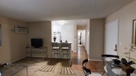 This single-floor residence offers a practical and open layout that seamlessly connects the main living areas. The living room is carpeted and provides a comfortable space anchored by a media console with a flat-screen television. Adjacent to the living area, a compact kitchen features white cabinetry, a full-size refrigerator, and a breakfast bar with two bar stools, creating a casual dining option. The kitchen's lighter flooring and flush ceiling light brighten the space, contrasting with the warmer tones of the surrounding rooms. A glass-top dining table with chairs is positioned nearby, offering a more formal dining setting. Hardwood flooring runs through the transitional areas and hallway, which extends toward additional rooms at the rear of the unit, including what appears to be a bathroom and further living spaces. The neutral taupe wall color throughout creates a cohesive, understated palette. A textured ceiling is consistent across the space. Overall, the layout is efficiently designed, with distinct functional zones flowing naturally from one to the next.