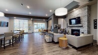 This well-appointed single-floor clubhouse offers a thoughtfully designed layout that blends comfort and functionality across a range of communal spaces. The grand entrance lobby sets an impressive tone with soaring ceilings, large floor-to-ceiling windows that flood the space with natural light, elegant tile flooring, a statement chandelier with gold accents, and a staffed reception counter. Sliding glass doors connect the lobby to the main lounge and social areas beyond.

The expansive main lounge features rich wood-style flooring, a stone-accented fireplace with a mounted flat-screen television, and generous seating arrangements including plush sofas and accent chairs. Large windows and glass doors along the exterior walls provide abundant natural light and direct access to outdoor areas. A dedicated game room offers a round game table, wall-mounted television, and natural light through wide windows.

The full kitchen is equipped with crisp white cabinetry, dark granite countertops, a white subway tile backsplash, a stainless steel sink, and a professional coffee station. A kitchen island with bar seating connects seamlessly to the lounge area, creating an ideal space for casual dining and entertaining. Throughout the clubhouse, modern drum pendant lighting, curated artwork, and contemporary furnishings contribute to a cohesive, upscale aesthetic.