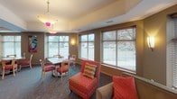 This community room is a thoughtfully designed, single-level shared space that offers a variety of functional areas to accommodate different activities and social settings. The room is finished with warm, earthy tones — predominantly tan and brown walls accented by bold pops of color from vibrant artwork and patterned upholstery — creating an inviting and lively atmosphere throughout.

A central lounge area anchors the space, featuring two generous sofas arranged around a fireplace, ideal for relaxed conversation. A colorful canvas artwork above the mantle adds a dynamic focal point. Pendant lighting with distinctive pink accents provides a warm glow overhead.

Multiple dining areas are arranged throughout the room, offering both square and round tables paired with wood-frame chairs featuring red patterned seat cushions. These areas are well-lit by natural light streaming through numerous large windows fitted with horizontal blinds, offering views of the surrounding outdoor environment.

A windowed nook provides additional flexible seating with accent chairs and a small table, creating a quieter retreat within the larger room. A dedicated kitchenette area, complete with cabinetry, countertops, a sink, and a water dispenser, supports catering and refreshment needs. The patterned carpet and tile flooring delineate the various zones within the space.