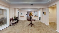 This single-floor senior living community is thoughtfully designed to provide a comfortable and functional environment for its residents. The main entrance opens into a welcoming lobby adorned with a decorative pendant light fixture, tray ceiling, patterned carpeting, and a central round pedestal table topped with a vibrant floral arrangement. A community bulletin board and elevator are conveniently located nearby, making navigation easy for all residents.

The spacious dining room is the heart of the community, featuring multiple dark wood dining tables paired with cushioned chairs, recessed lighting, and tray ceilings that lend an air of elegance. Burgundy valances frame windows that allow natural light to filter in and offer views of the surrounding greenery. Decorative wall art and floral accents contribute to the room's warm, inviting ambiance.

The facility includes two well-appointed kitchen areas. The primary kitchen is a galley-style space equipped with ample wooden cabinetry, white countertops, a full-size refrigerator, dishwasher, microwave, and coffee maker — providing everything needed for meal preparation. A secondary service kitchen features additional cabinetry, countertop workspace, and a utility sink, connecting directly to the dining area for efficient service. Throughout the space, warm peach-toned walls, light flooring, and tasteful décor create a cohesive, residential atmosphere.