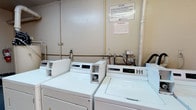 This shared laundry room offers a practical and straightforward space designed to meet residents' everyday laundry needs. The room features a row of commercial-grade Maytag top-load washing machines, each equipped with digital displays and coin-operated controls, ensuring reliable and efficient performance. The machines are positioned along the wall, with visible plumbing connections and utility piping that reflect the room's functional, no-frills design. The walls are finished in a neutral tone, and informational signage is posted prominently, providing users with important guidelines regarding accepted garments and usage policies, as well as emergency contact information. A warning notice is also displayed, reminding users of their responsibilities while using the facility. The layout is straightforward, allowing easy access to each machine. The utilitarian nature of the space prioritizes function over form, making it a dependable communal resource. Overall, the laundry room is a well-organized, essential shared amenity that supports the day-to-day convenience of its users.