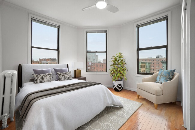 Bright and airy bedroom with three large windows, offering stunning city views, stylish decor, and a comfortable lounge chair. - Atrio Boston