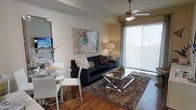This single-floor residence showcases an open-concept layout that seamlessly integrates the living and dining areas into one cohesive space. The rich hardwood flooring throughout anchors the room with warmth and character, complementing the neutral-toned walls that serve as a versatile backdrop for décor.

The living area is centered around a deep navy sofa, accented with decorative throw pillows, and paired with a sleek glass-and-chrome coffee table resting atop a plush, textured area rug. A large, colorful abstract painting adds a vibrant focal point to the main wall, while a warm table lamp provides ambient lighting.

A ceiling fan with integrated lighting is positioned centrally overhead, contributing to both the comfort and functionality of the space. Large sliding glass doors along one wall are dressed with sheer roller shades, allowing generous natural light to filter in while maintaining privacy.

The dining area features a round glass-top table surrounded by contemporary white chairs, creating an elegant yet casual setting ideal for everyday use. A wall-mounted mirror near the dining space enhances the sense of openness throughout the room.