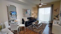 This single-floor residence showcases an open-concept layout that seamlessly integrates the living and dining areas into one cohesive space. The rich hardwood flooring throughout anchors the room with warmth and character, complementing the neutral-toned walls that serve as a versatile backdrop for décor.

The living area is centered around a deep navy sofa, accented with decorative throw pillows, and paired with a sleek glass-and-chrome coffee table resting atop a plush, textured area rug. A large, colorful abstract painting adds a vibrant focal point to the main wall, while a warm table lamp provides ambient lighting.

A ceiling fan with integrated lighting is positioned centrally overhead, contributing to both the comfort and functionality of the space. Large sliding glass doors along one wall are dressed with sheer roller shades, allowing generous natural light to filter in while maintaining privacy.

The dining area features a round glass-top table surrounded by contemporary white chairs, creating an elegant yet casual setting ideal for everyday use. A wall-mounted mirror near the dining space enhances the sense of openness throughout the room.