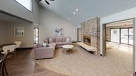 This single-level community room is thoughtfully designed to accommodate a variety of social gatherings and everyday use. The most striking feature is the expansive lounge area, which boasts soaring vaulted ceilings accented by a ceiling fan and recessed lighting. A full-height brick fireplace with a raised hearth and glass doors serves as the room's focal point, lending warmth and character to the space. Comfortable sectional seating and a round coffee table create an inviting atmosphere, while natural light filters in through nearby windows.

Adjacent to the lounge, a well-appointed kitchen provides ample functionality with dark countertops, white cabinetry, a stainless steel dishwasher, a microwave, and generous prep space along an L-shaped layout. A large window panel brings in additional natural light and connects the kitchen visually to the surrounding area.

A separate dining and activity zone features wood-look flooring, a wall-mounted flat-screen TV, and round tables with modern white chairs, making it ideal for meetings or casual dining. Track lighting overhead provides focused illumination throughout. Together, these distinct areas create a versatile, well-equipped community space suited for a range of social and functional purposes.