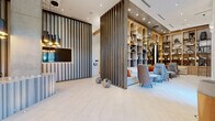 Located at 4401 Barclay Downs Dr in Charlotte, NC, this elegantly designed single-floor lobby and amenity space delivers a refined, hotel-inspired experience. The entrance greets visitors through striking double glass doors framed by floor-to-ceiling windows that flood the space with natural light, flanked by sculptural wood pedestals. Soaring ceilings amplify the sense of openness throughout.

The reception area features a textured, striated front desk paired with a dramatic slatted wood accent wall and a wall-mounted display screen. Vertical wood slat partitions serve as stylish architectural dividers that define zones while preserving an open, flowing layout.

A standout lounge area centers around a full-height built-in shelving wall crafted in warm wood tones, accessorized with books, art objects, and décor. Comfortable seating in neutral tones with orange accent pillows creates an inviting gathering space, complete with a rolling library ladder for added charm.

A dedicated meeting and collaboration area features a large communal table beneath a bold, sculptural copper chandelier, surrounded by floor-to-ceiling windows offering outdoor views. Nearby, a resident amenity bar includes a coffee station, under-counter refrigerator, and wall-mounted TV beneath another striking suspended art installation. The hallways are bright and well-appointed, with live-edge wood benches and warm accent lighting completing the polished aesthetic.