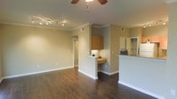 Located at 1420 McDermott Dr. in Allen, Texas, this apartment at Settler's Gate offers a well-designed single-floor layout that balances comfort and functionality. The open-concept living area is anchored by rich, dark hardwood-style flooring that extends throughout the main living space, creating a warm and cohesive aesthetic. Crown molding adds a refined architectural touch along the ceilings, which are outfitted with both a ceiling fan and a track lighting system, providing versatile illumination options for the room.

The living area flows naturally into the kitchen, which is separated by a half-wall breakfast bar — ideal for casual dining or entertaining. The kitchen features warm wood-toned cabinetry and a convenient built-in desk or counter nook, adding a practical workspace within the unit. A pass-through design keeps the kitchen connected to the living space while maintaining a sense of definition between the two areas.

Neutral, warm-toned walls throughout complement the flooring and cabinetry, contributing to an inviting atmosphere. A hallway visible from the main living area leads to the additional rooms of the unit. Built-in shelving is also visible, offering convenient storage solutions. Overall, the space presents a clean, functional, and comfortable living environment in a well-appointed Allen, TX community.