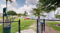 Located at 1410 Nottingham St in Huntsville, Texas, this outdoor amenity space is part of a multi-story residential community finished in a distinctive gray and white exterior. The grounds are thoughtfully designed to offer residents a variety of outdoor recreational areas set against a backdrop of mature pine and shade trees.

A standout feature of the property is the dedicated dog park, enclosed by black chain-link fencing for safety. The dog park includes canine play equipment, such as a red tunnel obstacle, providing an engaging environment for pets of all sizes. A paved walkway leads into the fenced area, making access convenient for residents.

Adjacent to the dog park is a well-maintained open green lawn, offering ample open space for outdoor activities. A charming gazebo structure with a pitched roof serves as a shaded gathering spot and is complemented by a blue bench nearby, ideal for relaxation. Additional mature trees throughout the grounds provide natural shade and a pleasant, park-like atmosphere.

The surrounding landscape features neatly trimmed grass, maintained pathways, and views of the broader neighborhood. Ample parking is visible along the perimeter of the property. The multi-story residential building anchors the space with its prominent architectural presence, featuring exterior stairwells and open-air corridors that connect the various levels of the community.