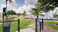 Located at 1410 Nottingham St in Huntsville, Texas, this outdoor amenity space is part of a multi-story residential community finished in a distinctive gray and white exterior. The grounds are thoughtfully designed to offer residents a variety of outdoor recreational areas set against a backdrop of mature pine and shade trees.

A standout feature of the property is the dedicated dog park, enclosed by black chain-link fencing for safety. The dog park includes canine play equipment, such as a red tunnel obstacle, providing an engaging environment for pets of all sizes. A paved walkway leads into the fenced area, making access convenient for residents.

Adjacent to the dog park is a well-maintained open green lawn, offering ample open space for outdoor activities. A charming gazebo structure with a pitched roof serves as a shaded gathering spot and is complemented by a blue bench nearby, ideal for relaxation. Additional mature trees throughout the grounds provide natural shade and a pleasant, park-like atmosphere.

The surrounding landscape features neatly trimmed grass, maintained pathways, and views of the broader neighborhood. Ample parking is visible along the perimeter of the property. The multi-story residential building anchors the space with its prominent architectural presence, featuring exterior stairwells and open-air corridors that connect the various levels of the community.