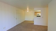 This single-floor unit offers a straightforward, functional layout centered around an open living area. The space features wall-to-wall carpeting in a neutral tone, creating a warm and cohesive feel throughout. A ceiling fan is centrally mounted overhead, providing both ventilation and a modest lighting fixture for the main living area. The walls are finished in a clean, off-white tone that keeps the space feeling bright and open.

A pass-through counter connects the living area to the kitchen, allowing for an open sightline between the two spaces. The kitchen is equipped with medium-toned wood cabinetry providing ample storage, a built-in microwave, and a dark range. The counter-height pass-through also serves as a casual dining or workspace option. A standard six-panel door near the living area provides access to either an entry point or a closet, adding practical utility to the layout. Overall, the unit presents a clean, move-in-ready environment with a simple and practical design well-suited for everyday living.