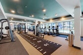 Located at 14965 W Encanto Blvd in Goodyear, Arizona, this generously sized fitness center offers a comprehensive, well-appointed exercise facility designed to accommodate a wide range of workout styles and fitness goals. The space is defined by its striking teal-painted tray ceilings, recessed lighting, and large windows that line the perimeter, flooding the interior with natural light and providing open views of the outdoor pool area and surrounding community.

The layout is thoughtfully organized into distinct zones. A central turf sprint track, marked with yardage lines, serves as a dynamic focal point for speed and agility training. Along the windowed wall, a row of treadmills, ellipticals, and stationary bikes — including an air bike — provides an extensive cardio section with outdoor views. A dedicated strength training area features multi-station cable machines, a functional trainer, and a cable pull system, while a free weight zone offers a full rack of dumbbells, adjustable benches, and a wall-length mirror with a mounted flat-screen display. Additional functional training equipment includes plyo boxes, a heavy bag, kettlebells, and medicine balls. Full-length mirrors throughout the space enhance the sense of depth and allow for form monitoring during workouts.