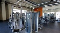 This single-level fitness center offers a comprehensive workout environment designed for both strength training and cardio exercise. The space is outfitted with a robust selection of Matrix-brand strength machines, including cable systems and resistance equipment, arranged to allow comfortable circulation between stations. Additional cardio machines are visible throughout the floor, providing a well-rounded range of training options.

Architecturally, the space features an industrial aesthetic with exposed ductwork, concrete ceilings, and pendant-style drop lighting that contributes to an open, loft-like atmosphere. Large windows along the walls invite natural light into the space, brightening the workout environment and creating an airy feel. An accent wall painted in a bold orange adds a dynamic visual element and energizes the overall ambiance.

The flooring is finished with dark rubber gym matting, offering durability and safety underfoot. The layout is thoughtfully organized, with equipment spaced to accommodate simultaneous use by multiple individuals. The combination of natural light, high exposed ceilings, and modern equipment makes this a functional and visually engaging fitness facility.
