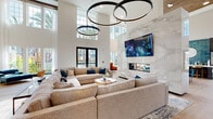 This expansive amenity lounge is defined by its dramatic double-height ceilings, creating an airy, grand atmosphere throughout the open-plan space. The centerpiece is a striking full-height marble accent wall housing a sleek linear fireplace and a large mounted television, serving as a visual anchor for the main lounge area. Oversized circular pendant lights suspended from above add a bold, modern design statement.

The main seating area features a generously proportioned sectional sofa in warm neutral tones, accented with teal and patterned throw pillows, arranged around a round wooden coffee table. Adjacent to the fireplace wall, a dining area offers a walnut oval table surrounded by plush teal velvet chairs, ideal for gatherings or collaborative use.

A sun-drenched reading nook lined with floor-to-ceiling windows floods the space with natural light and offers lush outdoor views of surrounding greenery and palm trees, furnished with cognac leather accent chairs and a metallic side table. Light hardwood flooring flows throughout, enhancing the bright, contemporary aesthetic.

The entry corridor features a built-in upholstered bench beneath a striking botanical-patterned wallcovering with warm pendant lighting, offering an inviting welcome. A reception desk area and decorative shelving complete the well-appointed space, making it functional, stylish, and comfortable.