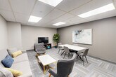 Located at 1400 Ouellette Ave in Windsor, Ontario, this thoughtfully designed single-floor space serves as a versatile lounge and meeting area with a compact kitchenette. The main room is divided into two functional zones: a comfortable lounge area and a dedicated meeting/dining area. The lounge features a light grey sofa accented with decorative pillows, two modern accent chairs with warm wood-toned legs, and a white-topped coffee table — all arranged beneath a round wall mirror that adds depth to the space. The meeting area offers a rectangular white table surrounded by upholstered grey chairs, ideal for small group gatherings or collaborative sessions. A wall-mounted flat-screen TV and a lush indoor plant add character and warmth to the room. A large abstract artwork in soft blues and whites serves as a focal point on the main wall. The space is finished with a patterned, multi-toned grey carpet tile floor and neutral grey walls, contributing to a clean, professional aesthetic. A drop ceiling with large recessed LED panel lights ensures bright, even illumination throughout. The adjoining kitchenette features white cabinetry, a stainless steel sink with granite-style countertops, and a full-size refrigerator, providing convenient amenities for extended use of the space.