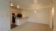 This single-story space showcases an open-concept layout that seamlessly connects the kitchen and living area, creating a functional and cohesive environment. The kitchen is equipped with black appliances, including a refrigerator, stove, and microwave, complemented by light wood cabinetry offering ample storage. A peninsula-style breakfast bar with a built-in sink serves as a natural divider between the kitchen and the adjoining living space, providing both additional prep space and casual seating opportunities. Track lighting installed above the kitchen ensures the workspace is well-lit, while a stylish pendant light hangs over the transition area, adding a decorative touch. The living area features neutral wall-to-wall carpeting that contributes to a warm and comfortable atmosphere, contrasting with the hard flooring found in the kitchen. The textured ceiling runs throughout, adding subtle visual interest. Neutral-toned walls throughout keep the space feeling bright and versatile, offering a clean canvas for personalization. The overall layout is efficient, making practical use of the available space while maintaining an open and airy feel.