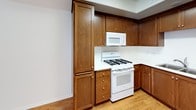 This single-floor residence offers a thoughtfully designed layout with a cohesive aesthetic throughout. The kitchen serves as a functional centerpiece, showcasing rich warm-toned wood cabinetry that extends from floor to ceiling, providing generous storage space. Light hardwood flooring anchors the kitchen, which is equipped with a gas range, over-the-range microwave, double basin sink, and a full-size refrigerator — all in white, creating a clean contrast against the wood cabinetry and bright countertops. A tray ceiling detail adds a subtle architectural touch to the space. The bedroom features neutral, wall-to-wall carpeting that adds warmth underfoot, smooth white walls, and a window fitted with horizontal blinds that allow for adjustable natural light. The room's simple, clean design provides a versatile canvas. The bathroom is finished with light tile flooring, a wood-toned vanity with a white countertop and integrated oval sink, a large frameless mirror, and a well-lit vanity fixture overhead. Built-in shelving is visible in the reflection, offering additional storage. Throughout the space, a consistent neutral color palette and practical finishes create a comfortable and functional living environment.